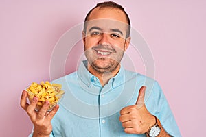 Young man holding bowl with macaroni pasta standing over isolated pink background happy with big smile doing ok sign, thumb up