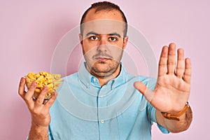 Young man holding bowl with macaroni pasta standing over isolated pink background with open hand doing stop sign with serious and