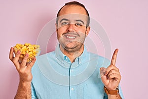 Young man holding bowl with macaroni pasta standing over isolated pink background surprised with an idea or question pointing