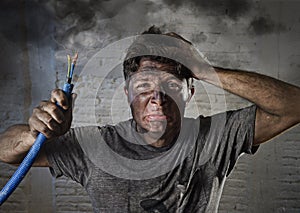 Young man holding cable smoking after electrical accident with dirty burnt face in funny sad expression