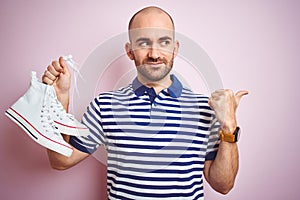 Young man holding casual sneakers shoes over pink isolated background pointing and showing with thumb up to the side with happy