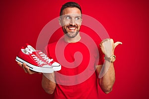 Young man holding casual sneakers shoes over red isolated background pointing and showing with thumb up to the side with happy