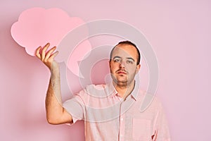 Young man holding cloud speech bubble standing over isolated pink background with a confident expression on smart face thinking