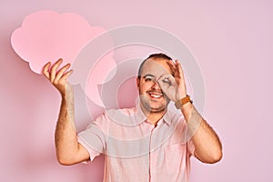 Young man holding cloud speech bubble standing over isolated pink background with happy face smiling doing ok sign with hand on