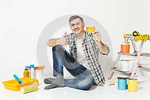 Young man holding credit card with money sitting on floor with instruments for renovation apartment isolated on white