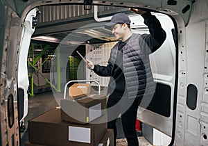 Young man holding a digital tablet while standing at van. Courier checking cardboard boxes on cart