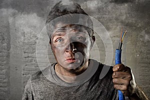 Young man holding electrical cable smoking after electrical accident with dirty burnt face in funny sad expression