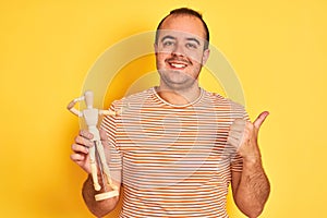 Young man holding figure of art dummy standing over isolated yellow background happy with big smile doing ok sign, thumb up with