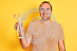 Young man holding figure of art dummy standing over isolated yellow background with a happy face standing and smiling with a