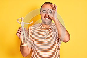 Young man holding figure of art dummy standing over isolated yellow background with happy face smiling doing ok sign with hand on