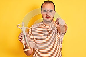 Young man holding figure of art dummy standing over isolated yellow background pointing with finger to the camera and to you, hand