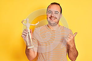 Young man holding figure of art dummy standing over isolated yellow background pointing and showing with thumb up to the side with