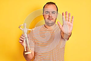 Young man holding figure of art dummy standing over  yellow background with open hand doing stop sign with serious and