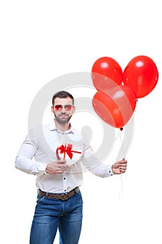 Young man holding gift box and balloons over white background. with glasses in the shape of heart. Looking at camera
