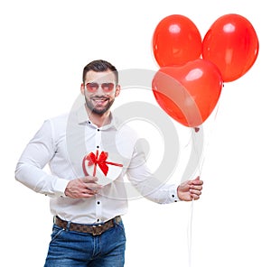 Young man holding gift box and balloons over white background. with glasses in the shape of heart. Looking at camera