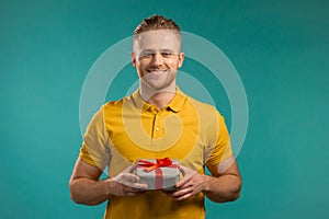 Young man holding gift box with bow, he wonders what is inside. Blue wall background. Guy smiling, he is happy to get