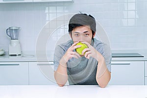 Young man holding green fresh apple breakfast in his kitchen looking camera. Handsome male eating fruit on