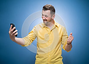 Young man holding in hands smartphone and making selfie while showing v-sign over blue background, dresses in yellow shirt. Trendy