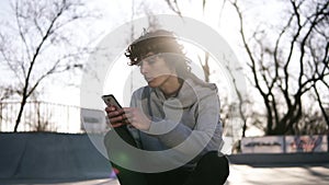 Young man is holding a mobile phone while sitting in a skate zone in skate park. A guy is surfing the web by the smart