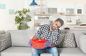 Young man holding plastic basin under water leakage from ceiling and calling plumber