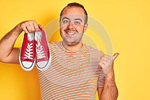 Young man holding red casual sneakers standing over isolated yellow background pointing and showing with thumb up to the side with