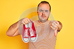 Young man holding red casual sneakers standing over  yellow background with angry face, negative sign showing dislike with