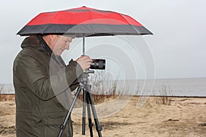 Young man is holding red umbrella and taking pictures of the beach at the rainy day