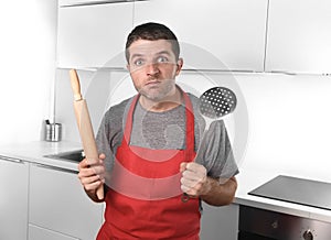 Young man holding skimmer and rolling pin wearing apron at home