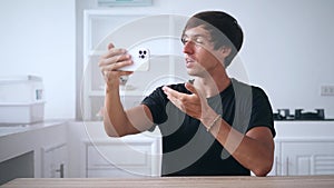 Young man holding smartphone and talking on a video call using his mobile phone, at home in the kitchen