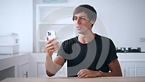 Young man holding smartphone and talking on a video call using his mobile phone, at home in the kitchen