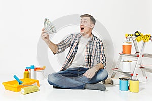 Young man holds bundle of dollars, cash money, sits on floor with instruments for renovation apartment isolated on white