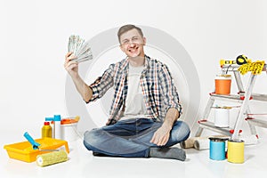 Young man holds bundle of dollars, cash money, sits on floor with instruments for renovation apartment isolated on white