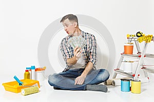 Young man holds bundle of dollars, cash money, sits on floor with instruments for renovation apartment isolated on white