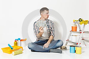 Young man holds bundle of dollars, cash money, sits on floor with instruments for renovation apartment isolated on white