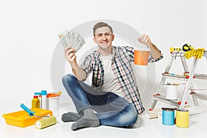 Young man holds bundle of dollars, cash money, sits on floor with paint can, instruments for renovation apartment