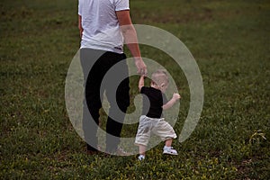 Young man holds the hand of little toddler. Father and son are walking along green field