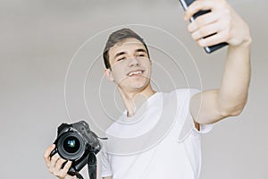 A young man holds a photo camera in his hand and makes a selfie by phone. Isolated gray background photo