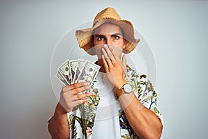 Young man on holidays holding dollars bank notes over white isolated background cover mouth with hand shocked with shame for