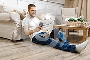 young man at home on the floor near the sofa working on a laptop with a phone