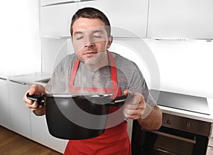 Young man at home kitchen in cook apron holding pot enjoying cooking smell