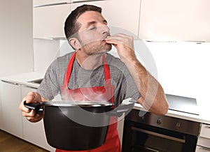 Young man at home kitchen in cook apron holding pot enjoying cooking taste
