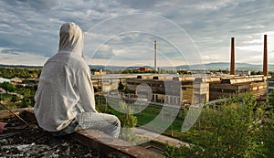 Young man in hood sitting and enjoy view to city Ceske Budejovice in sunset with old foundry, Czech republic