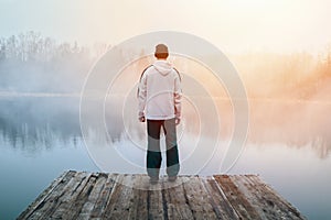 Young man in hoodie, hat and pants standing on wooden pier on pond shore with melancholy fog at sunrise. Czech morning landscape