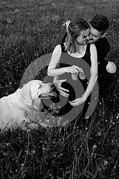 Young man hugs his pregnant wife, who is holding baby shoes in her hands, their smartest dog is lying next to her. Black and