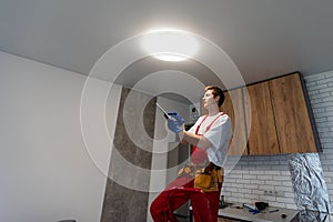Young man installing ceiling lamp on stepladder in kitchen