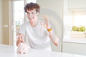 Young man investing money using piggy bank at home doing ok sign with fingers, excellent symbol