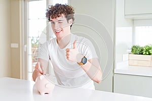 Young man investing money using piggy bank at home happy with big smile doing ok sign, thumb up with fingers, excellent sign