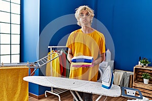 Young man ironing clothes at home puffing cheeks with funny face