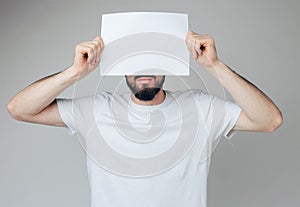 Young man isolated over background. Bearded guy cover part of face with white blank page. Hold it with both hands