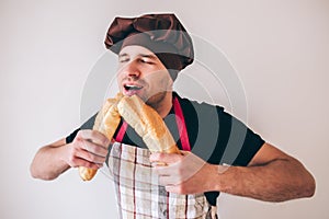 Young man  over background. Guy in cook uniform biting two pieces of french baguette and enoy it. Eating fresh photo